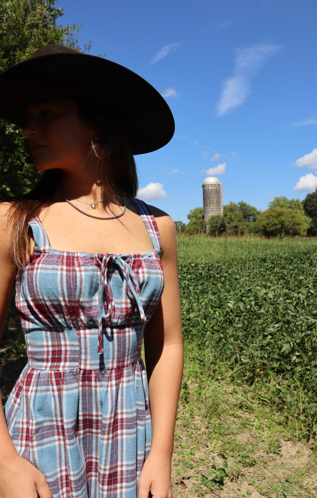 Person wearing a plaid dress and wide-brimmed hat standing in a field with a silo in the background.