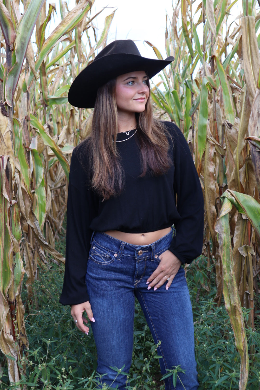 Person wearing a black coat, blue jeans, and a black hat standing in a cornfield.