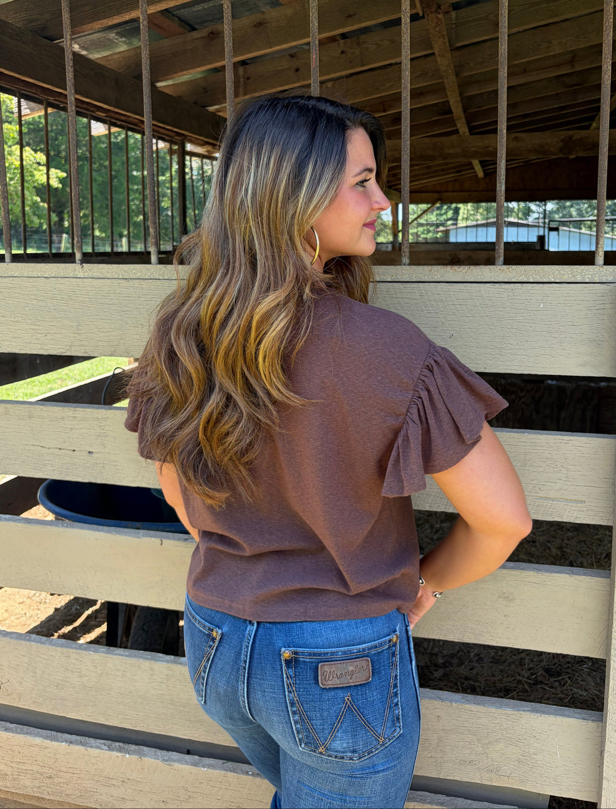 A woman with long brown and blonde highlighted hair stands in profile in front of a wooden stable, wearing a brown ruffle-sleeve top and blue jeans.