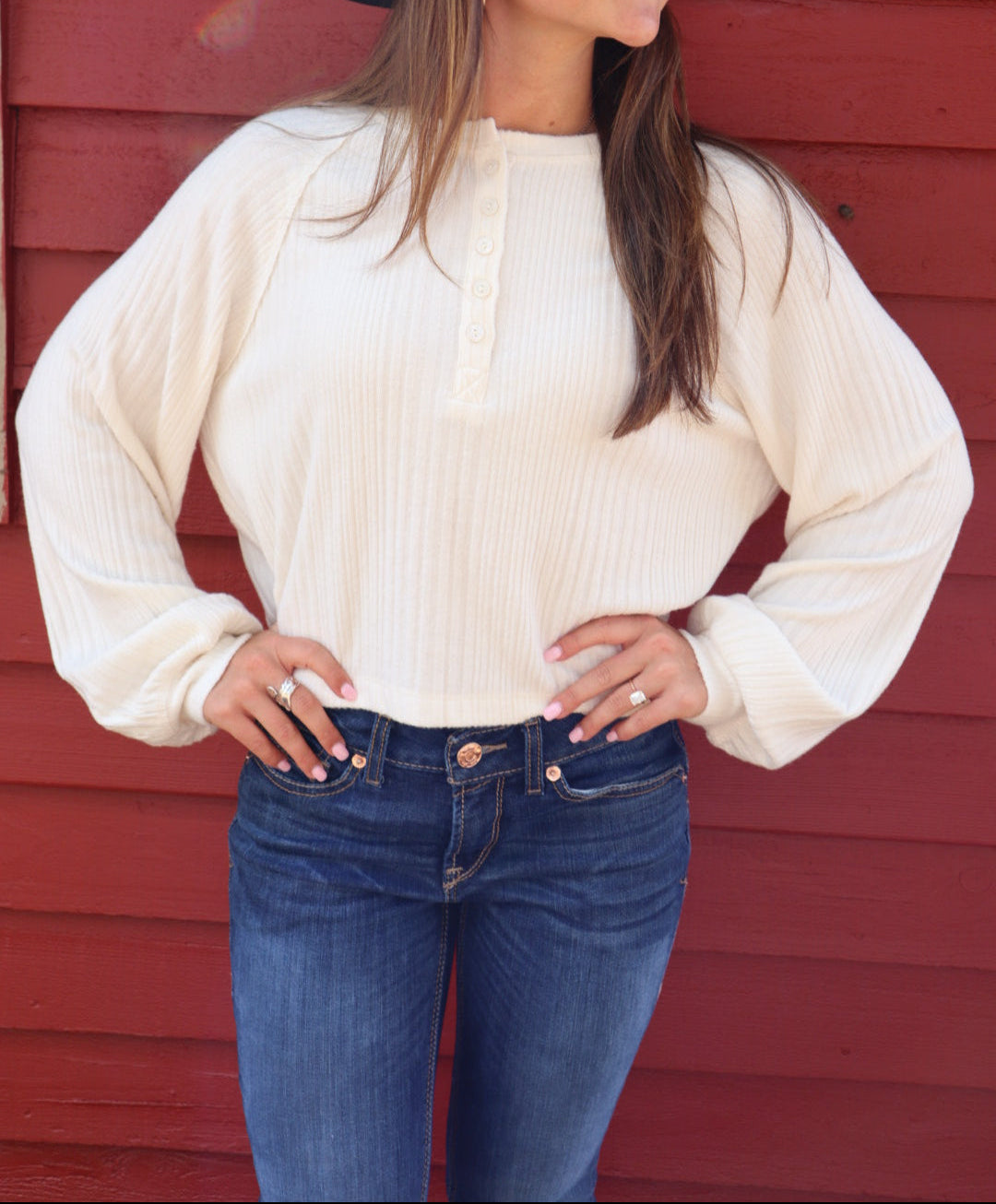 Woman wearing a white sweater, blue jeans, and a black hat against a red wooden wall.