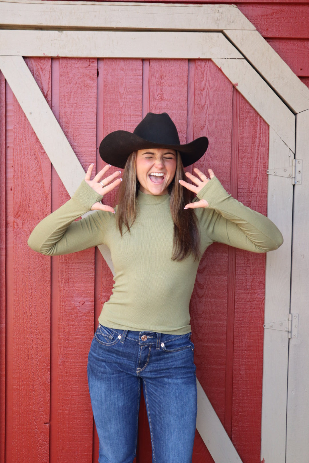 Person wearing a green sweater and blue jeans with a black hat against a red wooden background