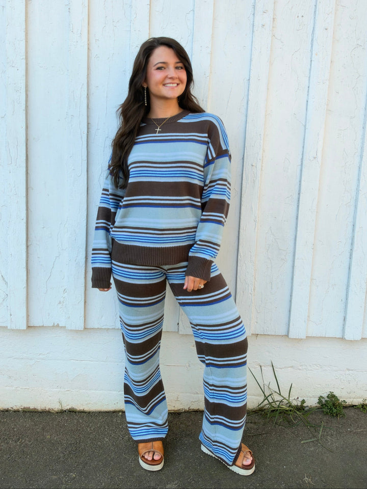 A woman stands in front of a white wooden wall wearing a matching long-sleeve top and wide-leg pants set with horizontal stripes in light blue, medium blue, brown, and white.