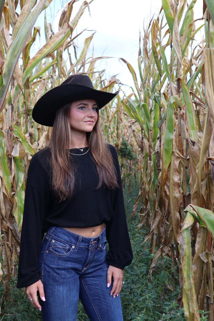 Person wearing a black top and jeans with a wide-brimmed hat standing among tall plants.