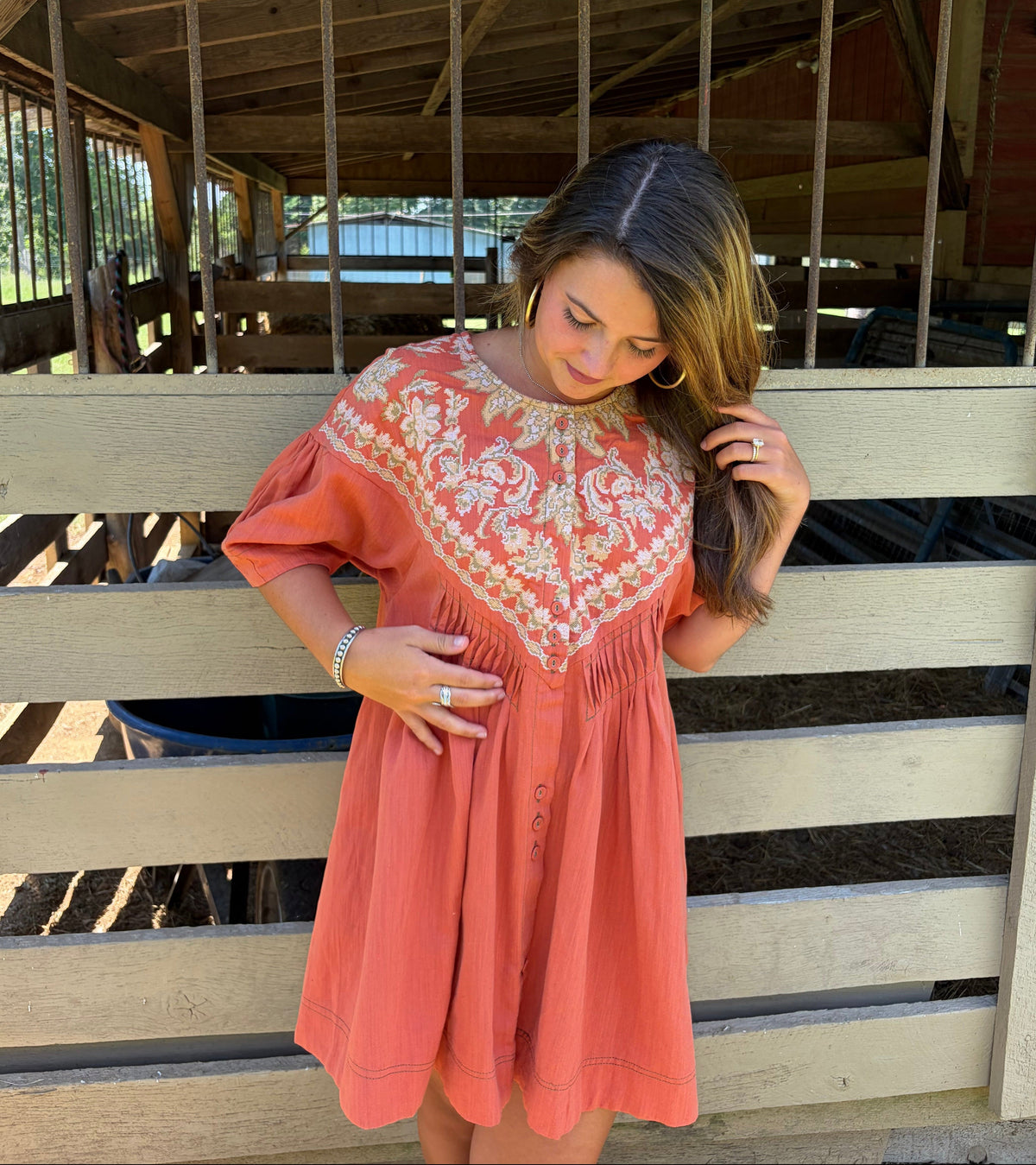 A woman wearing a coral pink and white embroidered dress stands in front of a wooden barn stall.